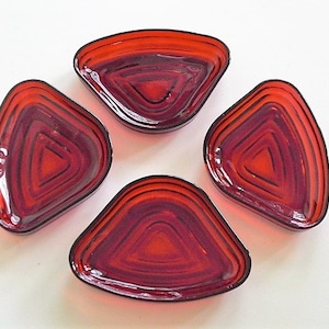 May include: Four red, triangular glass pieces with a layered design. Each piece has a dark red outline and a lighter red interior, creating a three-dimensional effect. The pieces are arranged in a circular pattern against a white background.