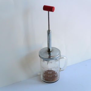 May include: A vintage glass measuring cup with a chrome lid and a red handle on a chrome plunger. The cup has markings for 1/4 cup, 1/2 cup, 3/4 cup, and 1 cup.