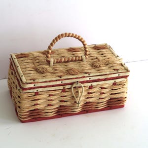 May include: A rectangular woven basket with a handle. The basket is made of light brown and red woven material. The handle is a twisted rope design. The basket has a red base and trim. The lid has a decorative clasp.