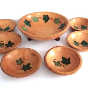 May include: A set of seven wooden bowls. The largest bowl is round with a dark brown base and green and light green ivy leaf design. Six smaller oval bowls have the same ivy leaf design. The bowls are light brown.