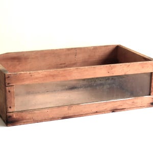 May include: A rectangular wooden box with a clear glass panel on one side. The box is made of light brown wood and has a handle cut out on one end. The box is likely vintage or antique.