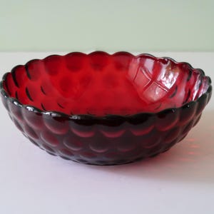May include: A ruby red glass bowl with a scalloped rim and a textured, bubble-like pattern on the exterior. The bowl is empty and sits on a white surface, with a light green background.