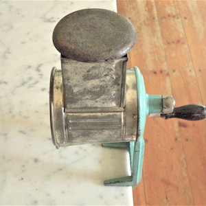 May include: An antique metal cheese grater with a wooden handle and a round, dark-colored top. The grater has a light blue handle and base, and is set against a marble surface and wooden floor.