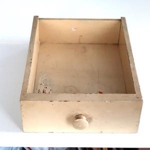 May include: A vintage, open wooden drawer with a round knob. The drawer is painted a faded cream color with visible wear and tear, including paint chips and discoloration, suggesting age and use. The interior shows signs of previous use.