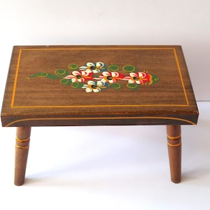 May include: A small, wooden step stool with a rectangular top and four short legs. The top is stained a dark brown and features a floral design in red, white, and green. The edges are trimmed with a gold border.
