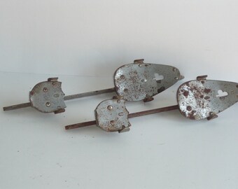May include: Four vintage metal ice skate blades with a distressed, rusty finish. Each blade has a rounded toe piece with a heart-shaped cutout and a rectangular heel piece. The blades are connected by a metal bar.