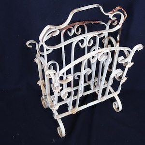 May include: An antique white metal magazine rack with a decorative, scrollwork design. The rack has a handle and is supported by curved legs. The metal shows signs of age with some rust.