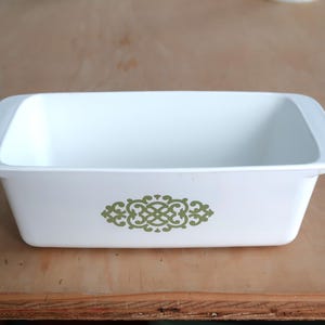 May include: A rectangular white baking dish with rounded edges and a decorative green floral design on the front. The dish is empty and sits on a light brown wooden surface.