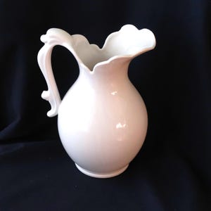 May include: A white ceramic pitcher featuring a rounded body, a wide pouring spout with a scalloped rim, and a curved, ornate handle. The surface of the pitcher is smooth and glossy, reflecting light across its curved form against a dark background.