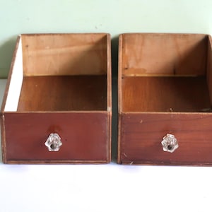 May include: Two wooden storage boxes with a dark brown front and a clear crystal knob. The boxes have a light brown interior and are set against a white and light green background. The boxes are empty.