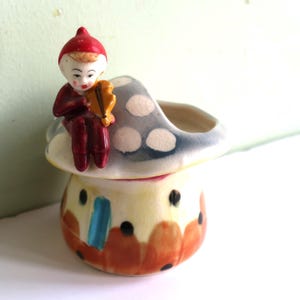 May include: A ceramic mushroom planter with a whimsical design. A small elf figurine sits on top of the mushroom, playing a musical instrument. The mushroom is painted with white polka dots and has a colorful base.