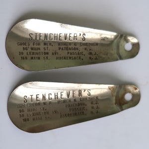 May include: Two vintage metal shoehorns with the name "Stenchever's" and store addresses printed on them. The shoehorns are oval-shaped with a hole at the top for hanging. The text indicates the store sold shoes for men, women, and children.