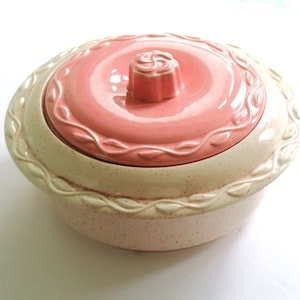 May include: A pink and white ceramic dish with a lid. The lid has a floral design and a knob in the center. The dish has a scalloped edge and a floral design around the rim.