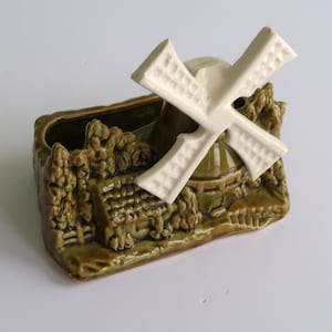 May include: A ceramic windmill planter in shades of olive green and cream. The rectangular planter features a detailed relief of a Dutch windmill scene, with a white windmill on top. The windmill blades are textured.