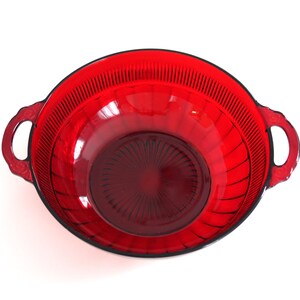 May include: A vibrant red glass bowl with two handles. The bowl has a ribbed design on the exterior and a circular pattern at the base. The bowl is likely for serving or display.