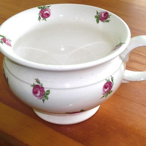 May include: White ceramic chamber pot with a handle and a wide rim. The pot is decorated with pink rose floral patterns. The handle is on the right side. The pot is sitting on a wooden surface.