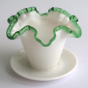 May include: A white glass vase with a ruffled rim, accented with a vibrant green trim. The vase sits on a matching white saucer, creating a delicate and elegant aesthetic. The design is reminiscent of vintage glassware.