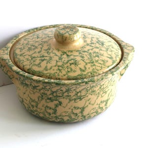 May include: A round, lidded ceramic casserole dish with a speckled green and beige pattern. The dish has two small handles and a knob on the lid. The overall design is vintage and rustic.