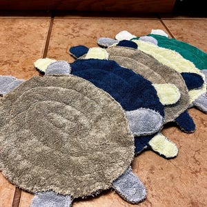 May include: A collection of four turtle-shaped fabric scrubbers arranged in a row. Each scrubber features a circular textured body in shades of tan, navy blue, light yellow, and dark green, with small light gray fabric appendages extending outward.