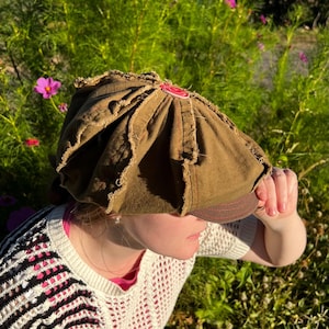 May include: An olive green newsboy cap featuring a multi-paneled construction with frayed, raw-edge seams. The hat has a structured brim with visible red stitching and a small, circular red emblem attached to the top center of the crown.