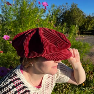 May include: A deep red corduroy newsboy cap featuring a structured, multi-paneled crown and a short, stiff brim. The fabric has a distinct vertical ribbed texture, and the hat is designed with a full, rounded shape that sits comfortably on the head.