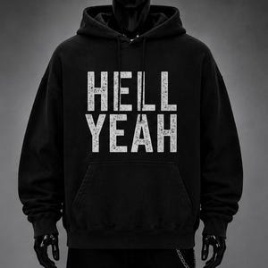May include: A black hooded sweatshirt featuring the text HELL YEAH printed in a distressed white font across the chest. The garment includes a front kangaroo pocket, long sleeves with ribbed cuffs, and a drawstring hood for a classic pullover design.