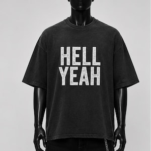 Könnte beinhalten: Ein anthrazitfarbenes Kurzarm-T-Shirt mit Rundhalsausschnitt und einem verwaschenen weißen Grafikdruck auf der Brust, der HELL YEAH in fetten Blockbuchstaben zeigt. Das Kleidungsstück hat eine lockere Passform mit überschnittenen Schultern.