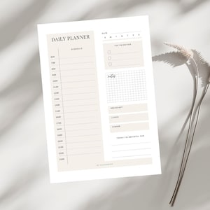 May include: A beige and white daily planner with the words "Daily Planner" at the top. The planner includes sections for scheduling, top priorities, notes, and meals. The time schedule is from 6:00 to 23:00. The planner is on a white surface.