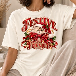 Op de afbeelding: Een off-white t-shirt met korte mouwen en een afbeelding van een norse kat met een rode kerstmuts en een grote rode strik. De tekst "FESTIVE" en "NOT FRIENDLY" is in het rood gedrukt met gouden accenten, omringd door hulstbladeren.