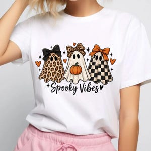 Könnte beinhalten: Ein weißes T-Shirt mit einer Grafik von drei Geistern mit Schleifen. Der erste Geist hat ein Leopardenmuster, der zweite ist schlicht mit einem Kürbis und der dritte hat ein schwarz-weißes Schachbrettmuster. Der Text Spooky Vibes steht darunter.