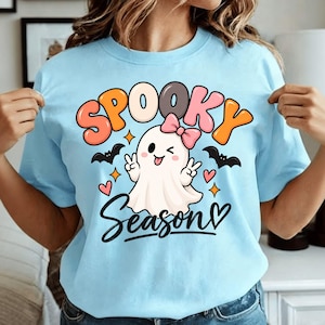 Può includere: Una t-shirt azzurra con la grafica di un fantasma bianco con un fiocco rosa, che fa l'occhiolino e il segno di vittoria. Il testo "SPOOKY Season" è scritto in lettere colorate, circondato da piccoli pipistrelli neri, cuori rosa e stelle.