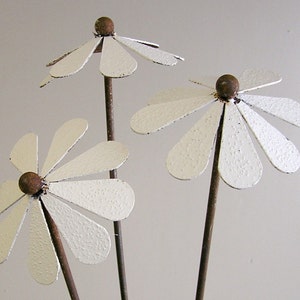 May include: Three metal daisy flowers with white petals and brown centers. The flowers are on thin, dark brown stems. The petals have a textured, slightly distressed appearance, suggesting a rustic or vintage style.
