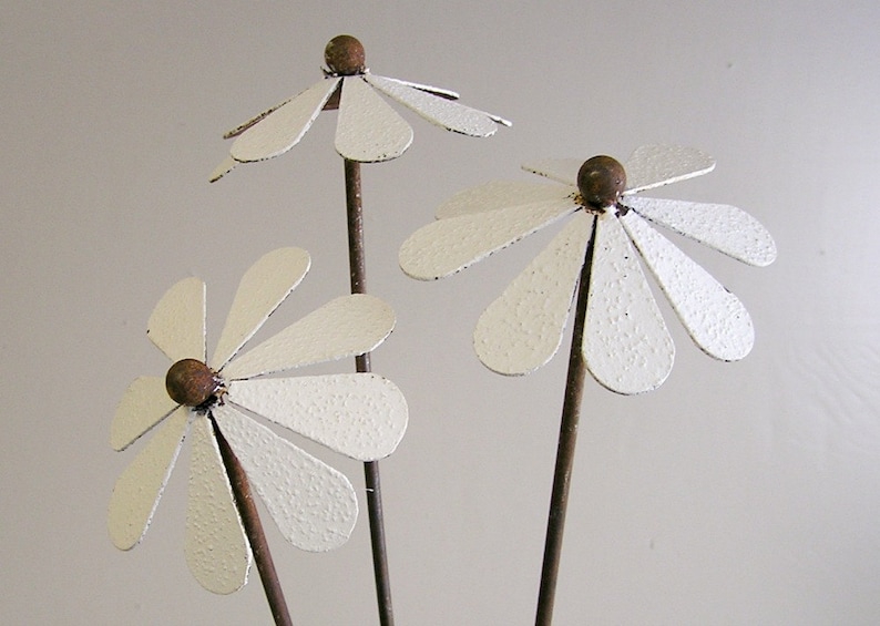 May include: Three metal daisy flowers with white petals and brown centers. The flowers are on thin, dark brown stems. The petals have a textured, slightly distressed appearance, suggesting a rustic or vintage style.