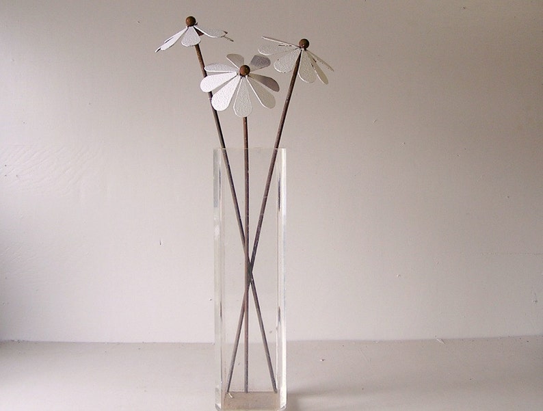 May include: A clear rectangular vase holds several artificial white daisy flowers with brown stems and wooden centers. The vase sits on a white surface against a white background. The flowers are a decorative accent.