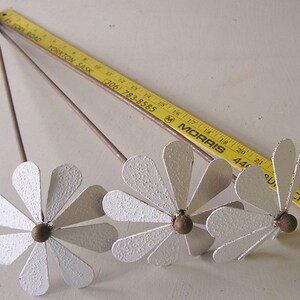 May include: Three metal daisy flower sculptures with white petals and brown centers. The flowers have long stems and are arranged on a white surface. A yellow ruler is in the background.