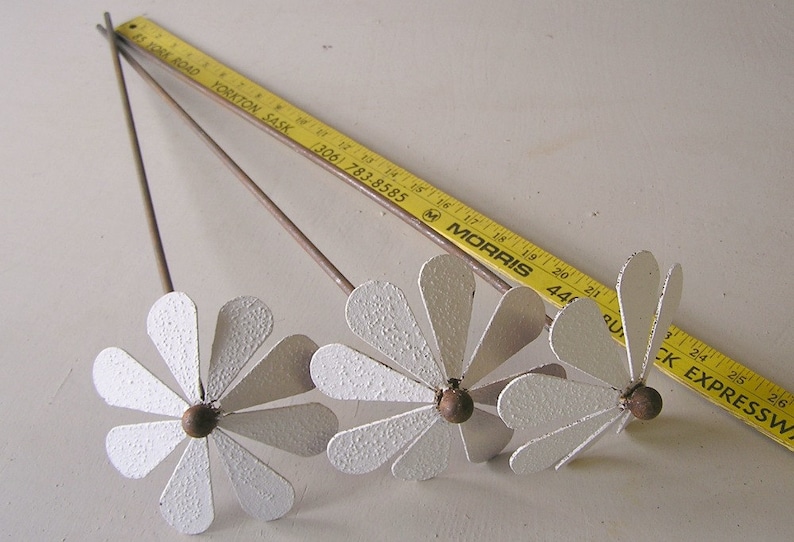 May include: Three metal daisy flower sculptures with white petals and brown centers. The flowers have long stems and are arranged on a white surface. A yellow ruler is in the background.