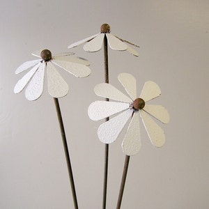 May include: Three metal daisy flower garden stakes. Each flower has white petals and a brown center. The stems are a dark brown color. The flowers are a decorative garden accent.