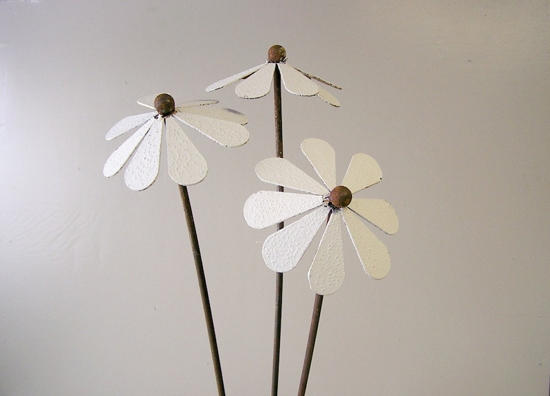 May include: Three metal daisy flower garden stakes. Each flower has white petals and a brown center. The stems are a dark brown color. The flowers are a decorative garden accent.