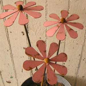 May include: Three pink metal flower garden stakes with brown centers and distressed edges. The flowers are in a galvanized metal pot against a white, weathered wooden background. The flower petals are a soft pink color.