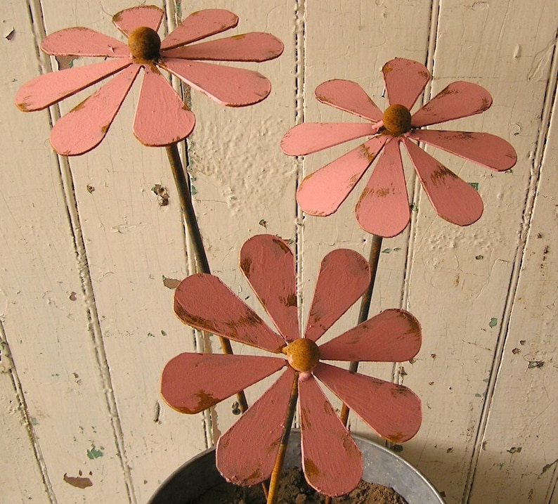 May include: Three pink metal flower garden stakes with brown centers and distressed edges. The flowers are in a galvanized metal pot against a white, weathered wooden background. The flower petals are a soft pink color.