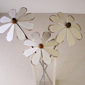 May include: Three metal daisy flowers with white petals and brown centers are arranged in a clear glass vase. The petals have a distressed, aged look with brown edges. The stems are thin and gray.