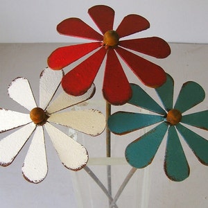 May include: Three metal daisy flowers in a vase. The flowers are painted red, white, and teal, with a distressed finish. Each flower has a yellow center and is on a metal stem. The vase is clear.