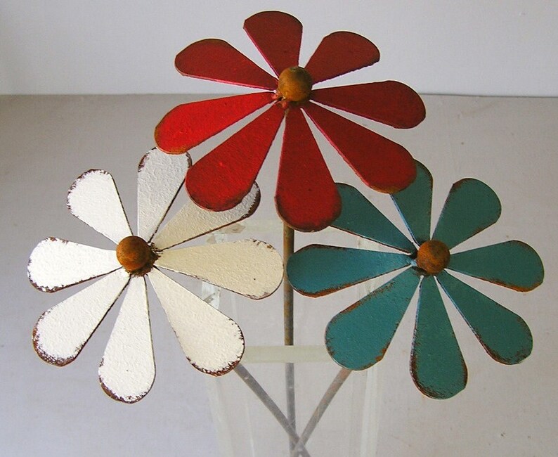 May include: Three metal daisy flowers in a vase. The flowers are painted red, white, and teal, with a distressed finish. Each flower has a yellow center and is on a metal stem. The vase is clear.