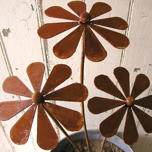 May include: Three rustic metal flower sculptures with a weathered, rusty brown finish. Each flower has a central sphere and petal-shaped leaves. The stems are thin and extend from a metal pot, suggesting garden decor.