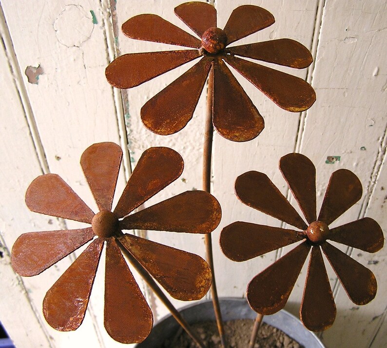 May include: Three rustic metal flower sculptures with a weathered, rusty brown finish. Each flower has a central sphere and petal-shaped leaves. The stems are thin and extend from a metal pot, suggesting garden decor.