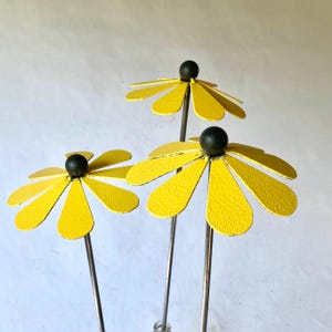 May include: Three metal daisy garden stakes. Each daisy has bright yellow petals and a black center. The stems are silver. The stakes are designed to be placed in a garden or flower bed.