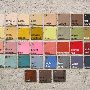 May include: A collection of painted color swatches in various shades, including white, yellow, pink, blue, green, and brown. Each square has a small hole for hanging and a label with the color name.