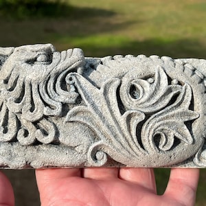 Może przedstawiać: Prostokątna, szara rzeźba w kamieniu z reliefem przedstawiającym szczegółową rzeźbę głowy mitycznego stworzenia i ozdobnych liści. Płaskorzeźba jest trzymana w dłoni, ukazując misterne detale i fakturę.