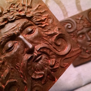May include: A close-up of a brown, textured, rectangular plaque with a detailed carving of a bearded man's face. The man's eyes are closed and his hair is flowing. The plaque is decorated with intricate carvings of seaweed and other sea-life.