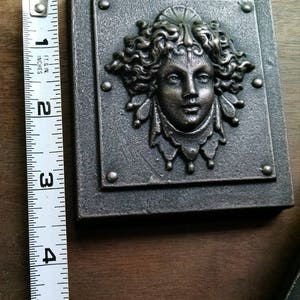 Pu&ograve; includere: Una placca da parete in metallo argentato con una scultura a rilievo di un viso di donna. La donna ha i capelli lunghi e ricci e indossa una corona. La placca &egrave; larga circa 10 cm e alta 12,5 cm.